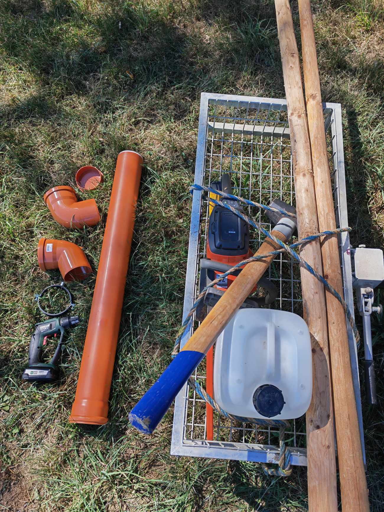 Material und Werkzeug für den Bau der Niederwildtränke: KG-Rohr, Bögen, Muffenstopfen, Schelle, Pflöcke und Werkzeug im Gras ausgelegt